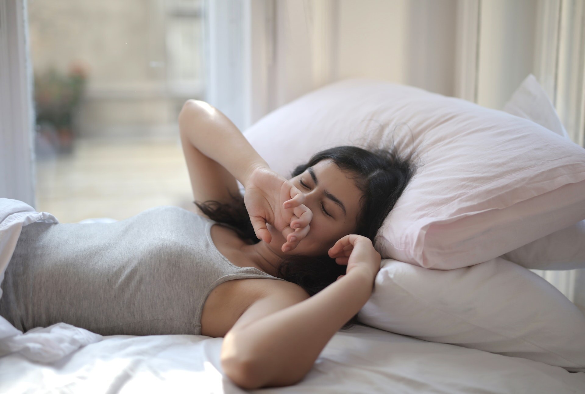 A woman with dark brown hair laying on a bed with closed eyes, her hand to her mouth as she yawns