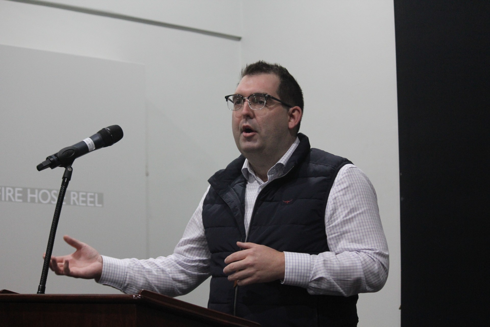 A dark-haired man in a dark vest stands at a lectern and speaks into a microphone.