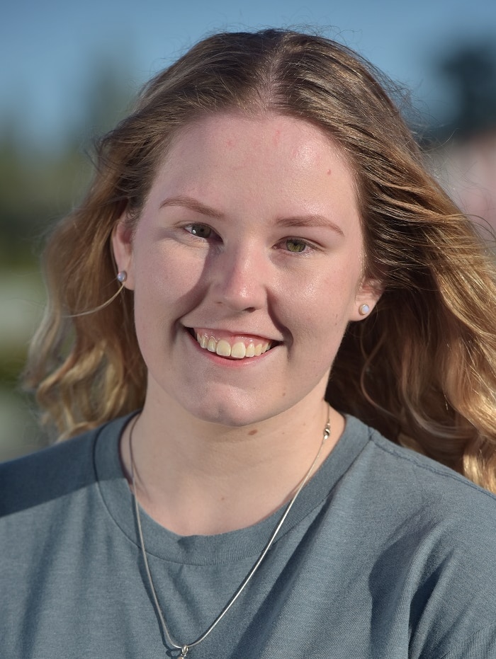 A portrait picture of a young woman with long, wavy blonde hair in a grey jumper, smiling.