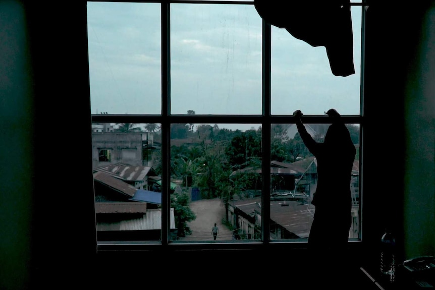 A victim of human trafficking from Myanmar's Kachin state stares out a window into a dirt street with rusted roofs.