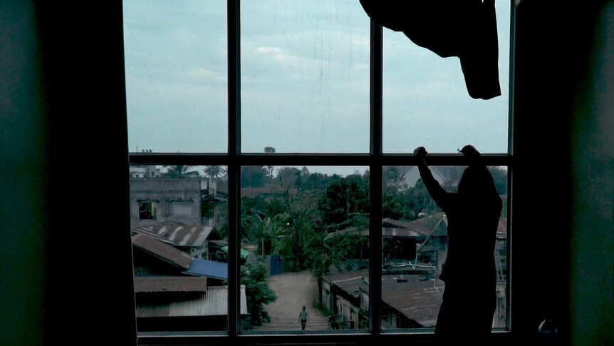 A victim of human trafficking from Myanmar's Kachin state stares out a window into a dirt street with rusted roofs.