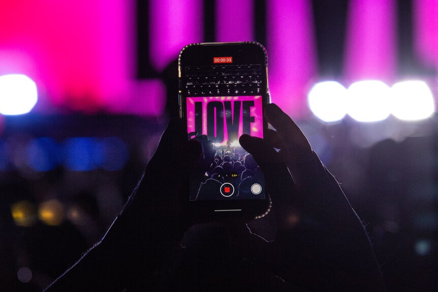 Hands in a crowd hold up a smartphone in video mode with the word LOVE visible on the screen