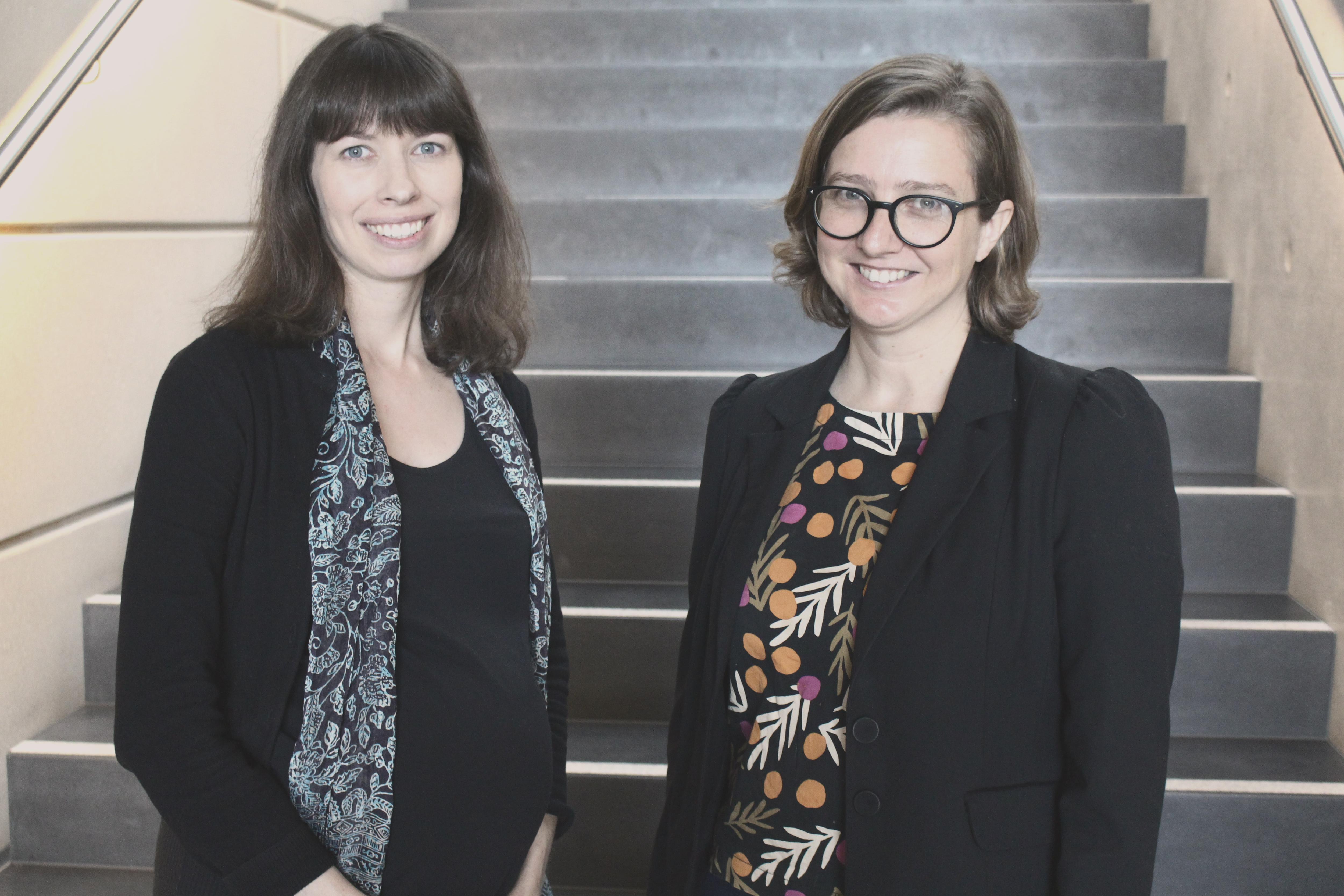 Two women smile at the camera, in front of a staircase.