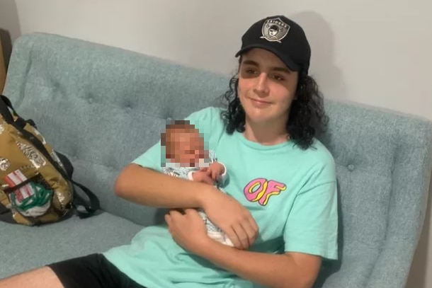 Young man with long curly hair wearing a black cap, sitting on a couch holding a newborn baby.