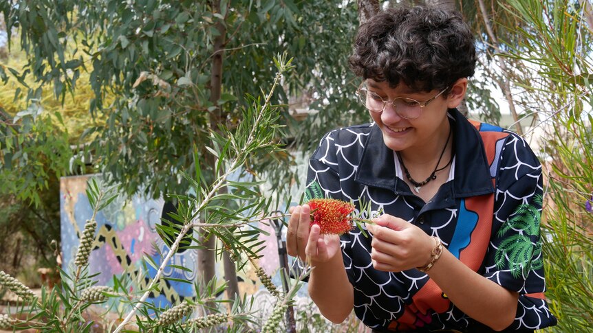 A smiling student with short dark curly hair wearing glasses and a colourful t-shirt holds a bottlebrush.