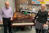 A man in his 80s and a woman in her 50s stand on either side of a grand piano.