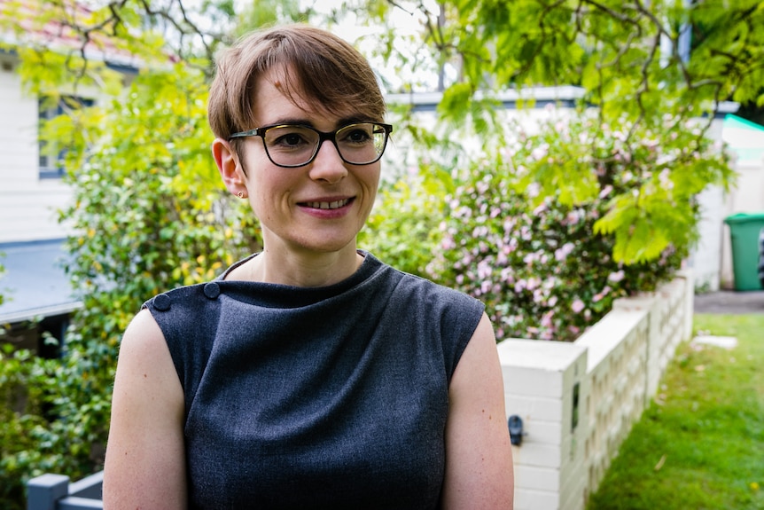 Woman in sleeveless blouse stands before a front garden.