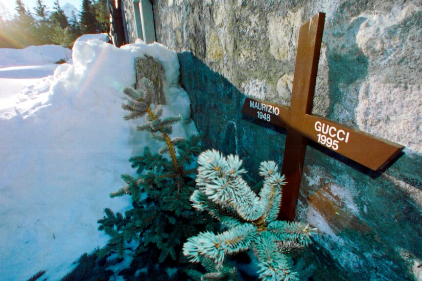 A wooden cross bearing the words MAURIZIO GUCCI 1948 1995 is propped up against a stone wall in the snow