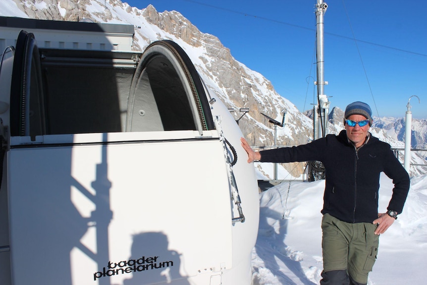 Dr Vogelmann in sunglasses and a beanie leaning against a rooftop telescope with one hand on it