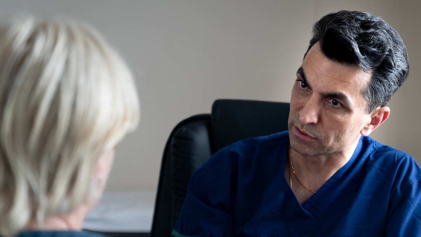 A male surgeon in scrubs gestures while explaining something to a blonde patient