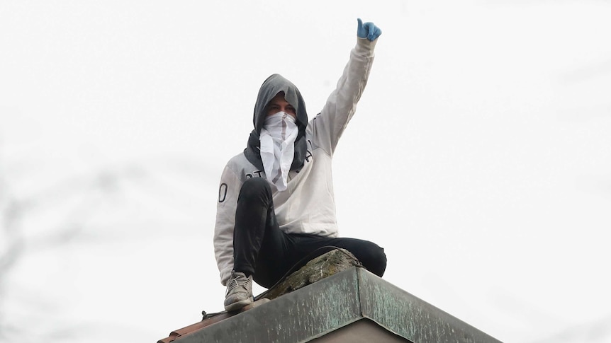 A man stands on the roof of a prison his face is covered and his arm is the air with his thumb up