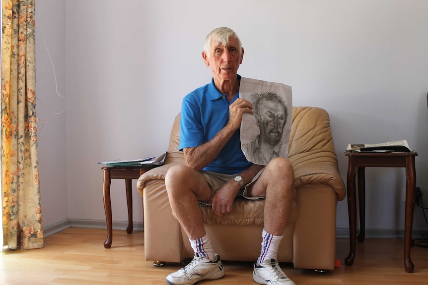 Kevin Handforth sits in armchair holding up sketch portrait of man