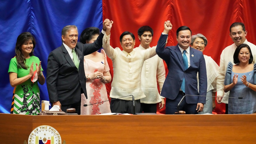 Three men link hands held up in air with relatives standing behind,