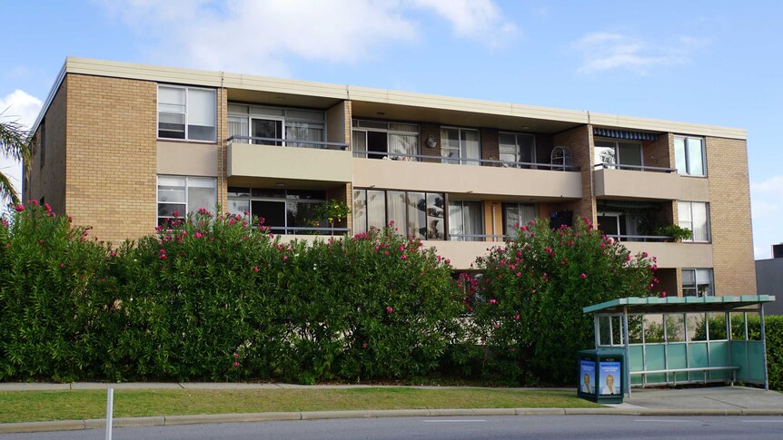 A wide shot of a block of apartments, with a hedge of bushes and a bus stop in the foreground.