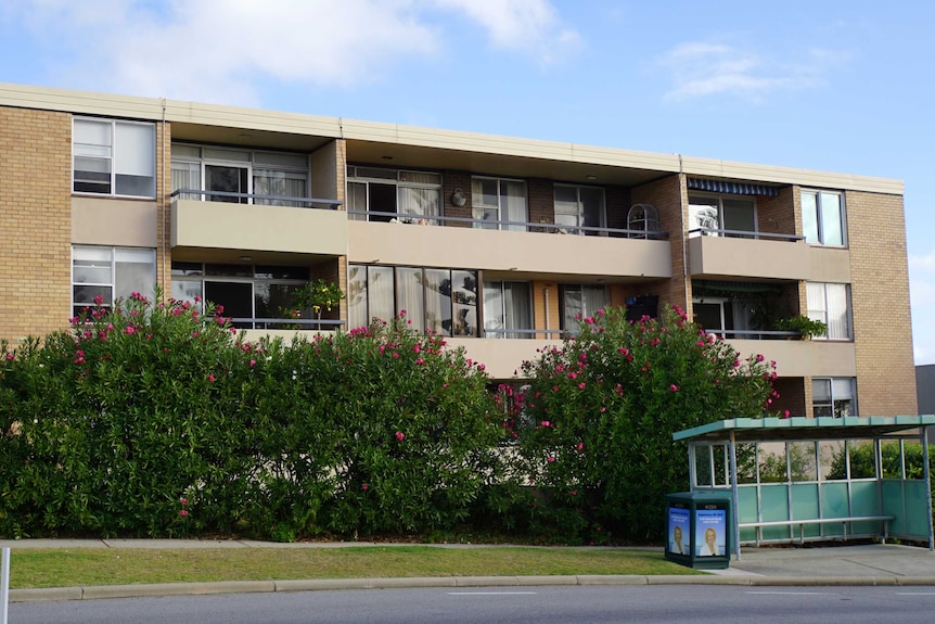 A wide shot of a block of apartments, with a hedge of bushes and a bus stop in the foreground.