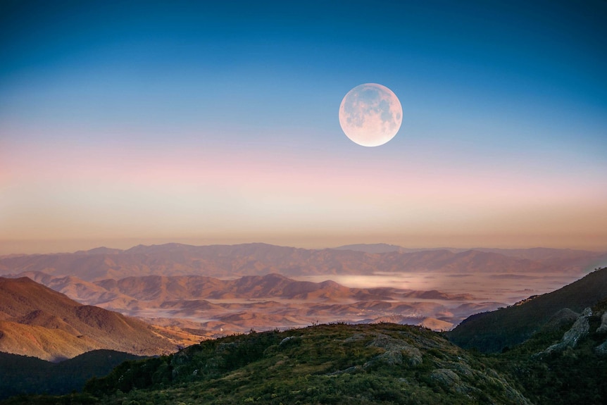 Moon in day sky over mountains.