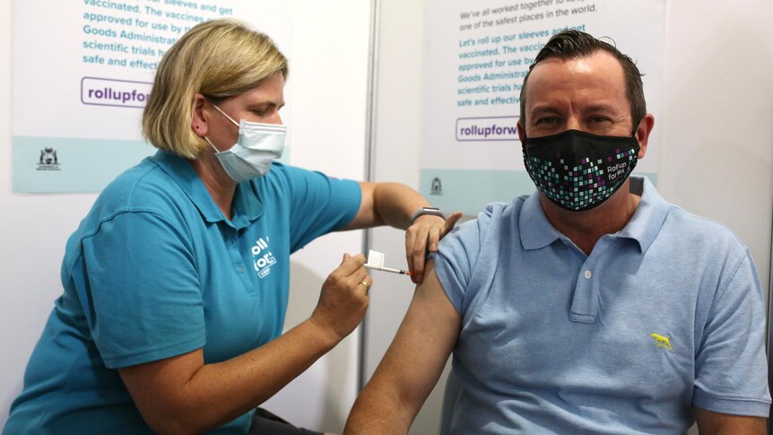 Mark McGowan smiles as he receives his jab from a blonde nurse with a blue shirt