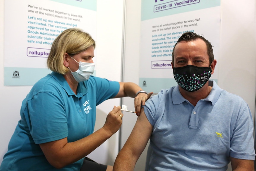 Mark McGowan smiles as he receives his jab from a blonde nurse with a blue shirt