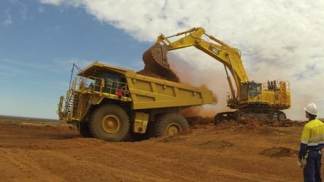 A digger dumps dirt into a haulpak as a worker in hi-vis clothing watches on.