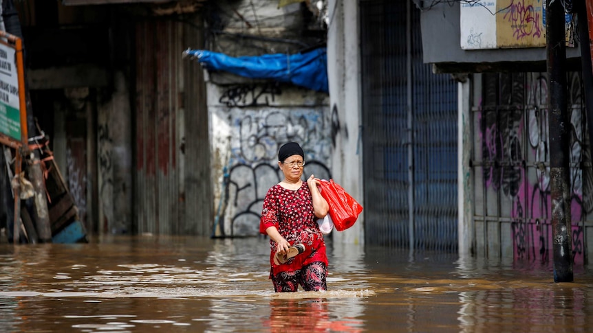 A woman wearing a red and black patterned outfit holds her shoes and plastic bag as she walks through water over knee-height