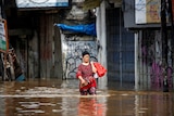 A woman wearing a red and black patterned outfit holds her shoes and plastic bag as she walks through water over knee-height
