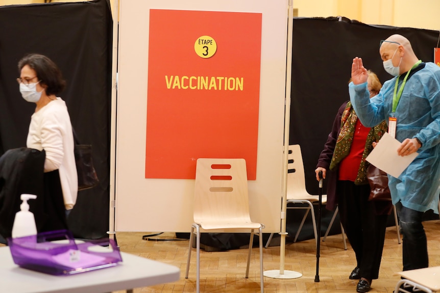 Un hombre vestido con matorrales señala a una mujer frente a un cartel que dice "El tercer paso, la vacunación.".