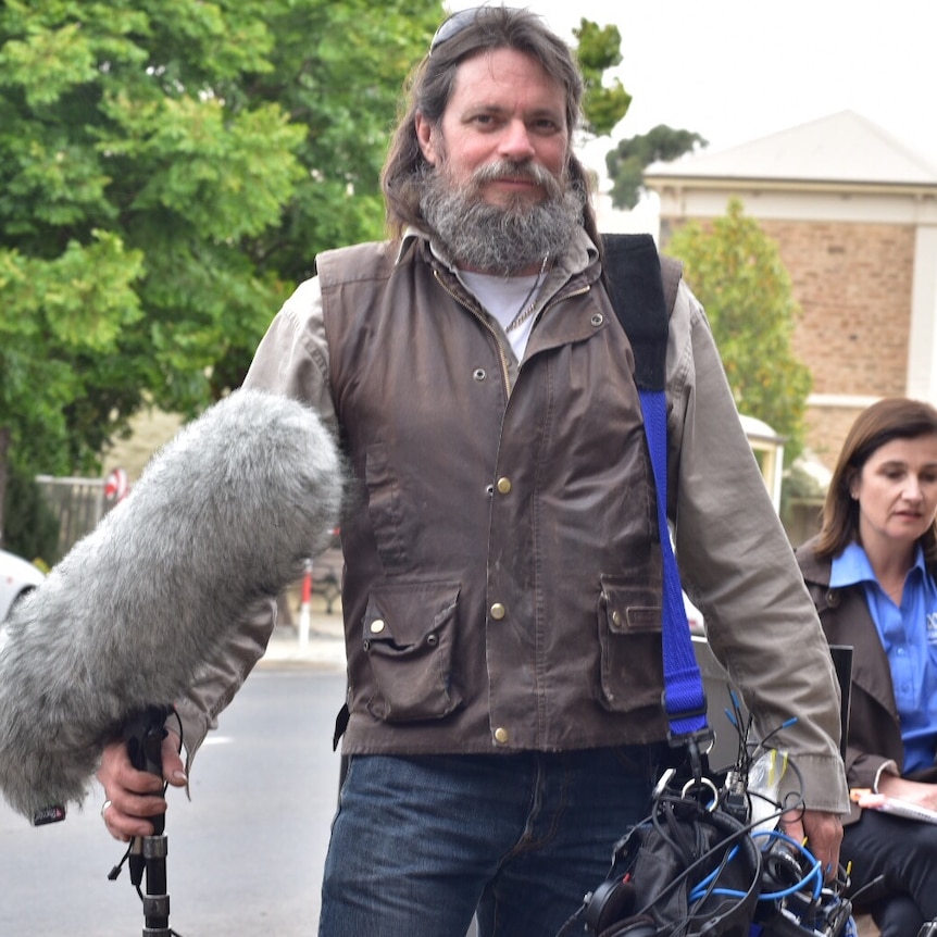 Tony Hill standing in Adelaide street holding audio equipment over his left shoulder and boom microphone in his right hand.