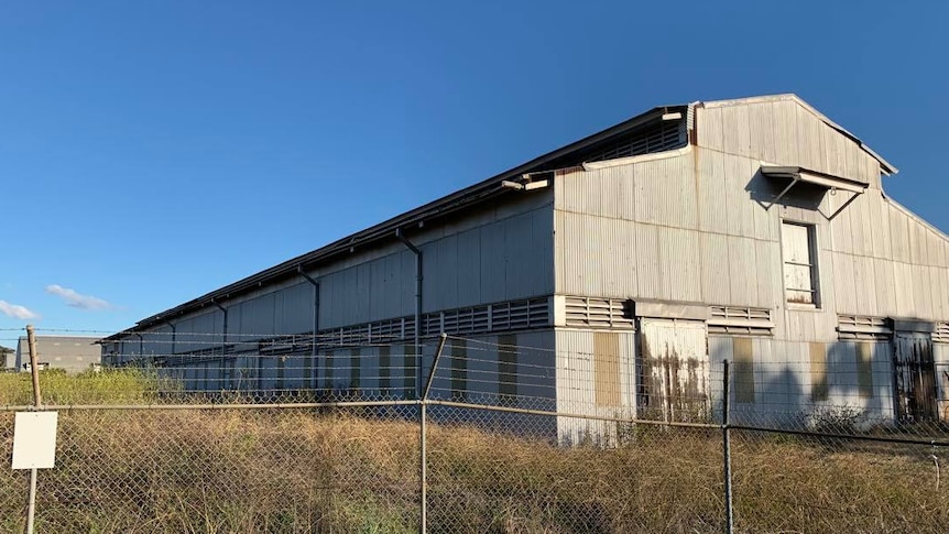A large industrial hangar amongst overgrown grasses with a  barbed wire fence in the foreground