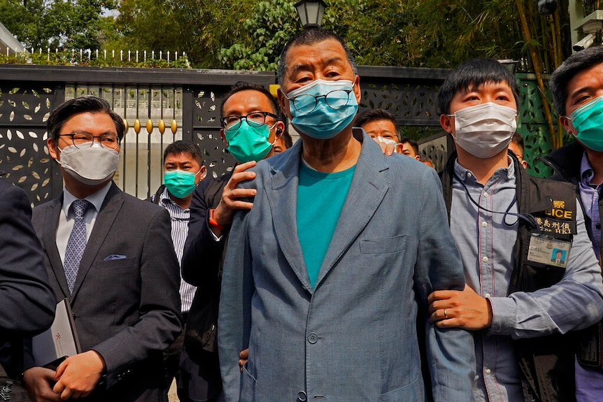 You see an elderly man in a grey suit and aqua t-shirt held by police in casual clothes who all wear face masks.