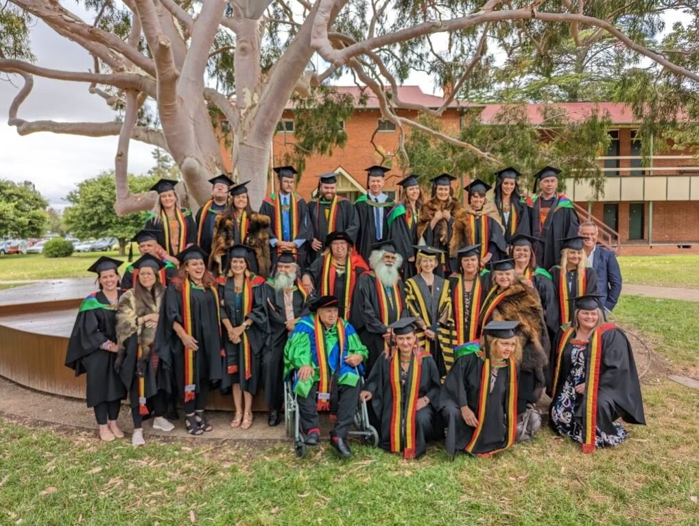 A group of university graduates stand under a tree in black caps and gowns. A man in a wheelchair in a green robe beside them
