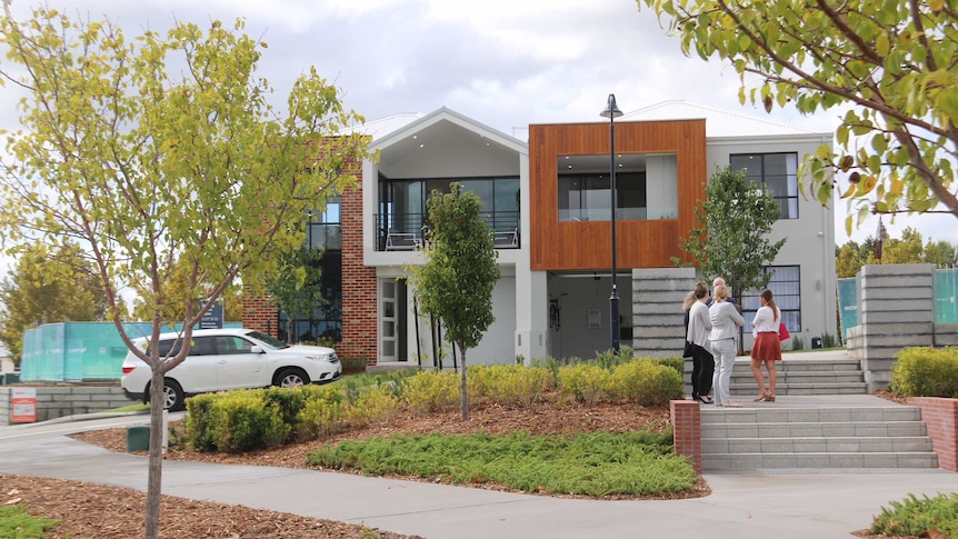 Several people stand outside a small, new two-storey micro lot display home with lots of gardens surrounding.