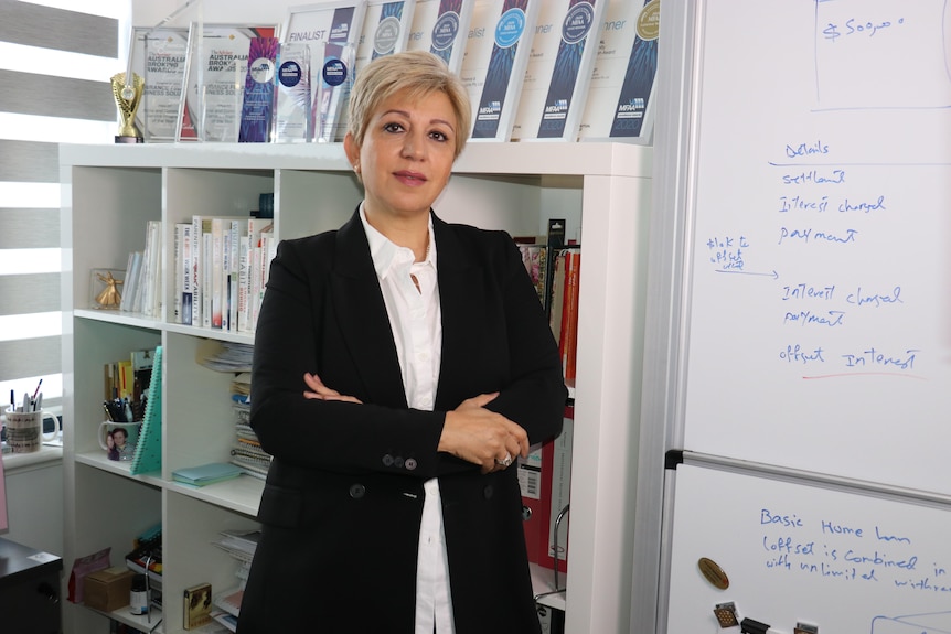 A middle aged woman wearing a black blazer and white shirt leans on a white bookcase with her arms crossed.