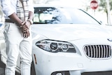 The bottom half of a man, standing in front of a white BMW, wearing a conspicuously expensive watch and scarf.