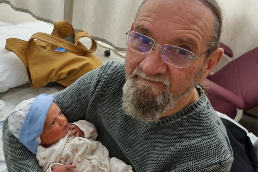 Gregory wearing glasses, smiling, holding baby William in his arms, baby's eyes open.