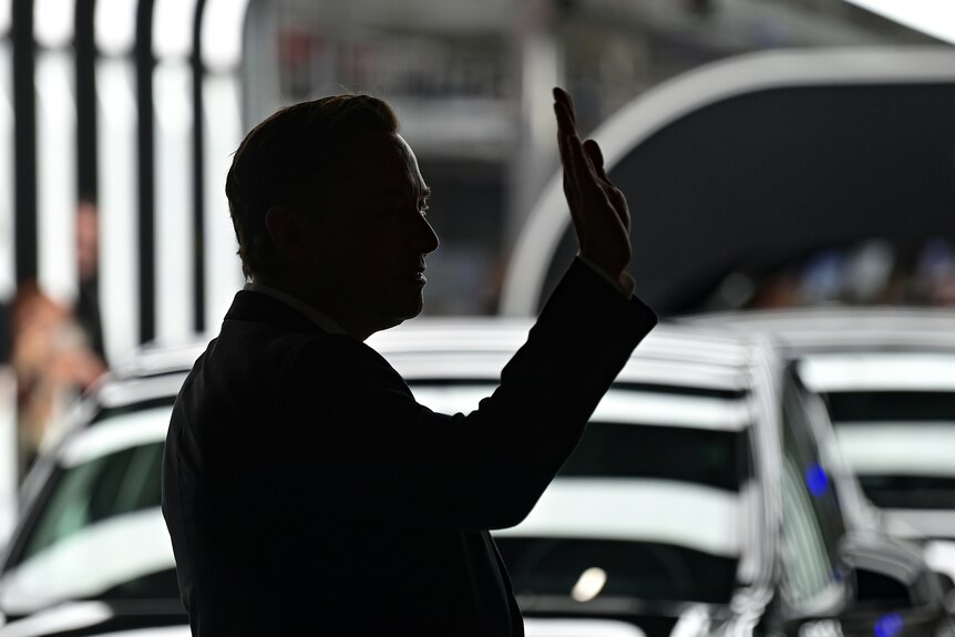 The silhouette of Elon Musk is visible, as he waves in front of a row of cars