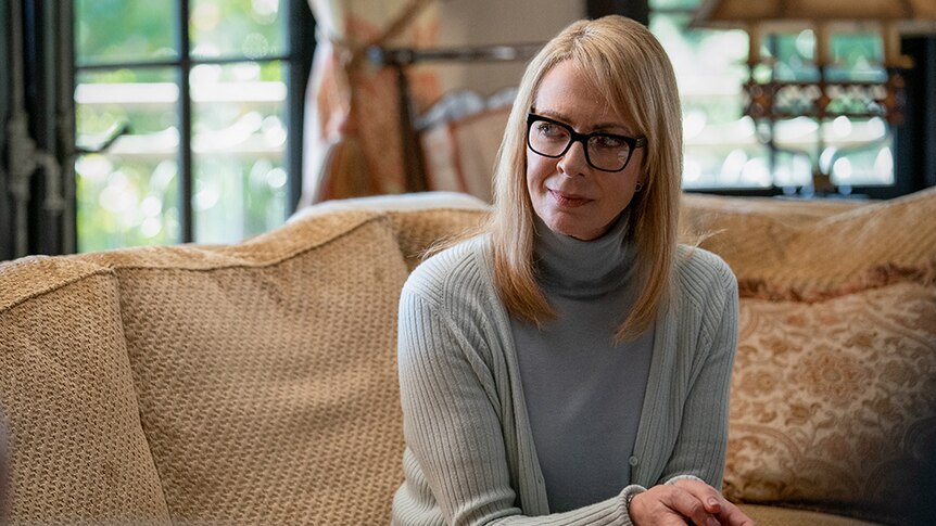 A woman with black square glasses and mid-length blonde hair wears gray cardigan and sits on couch in well lit living room.