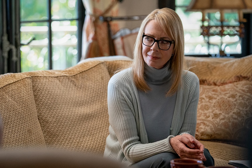 A woman with black square glasses and mid-length blonde hair wears gray cardigan and sits on couch in well lit living room.