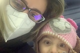 Kristy Colakidis wears a face mask on a plane while her young daughter smiles next to her.