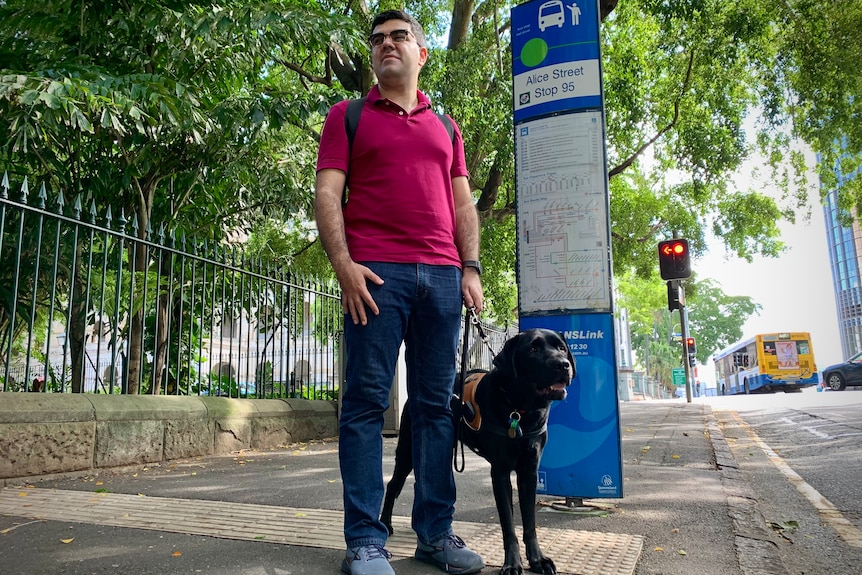 A man of Latin American background standing at a bus stop with a black guide dog