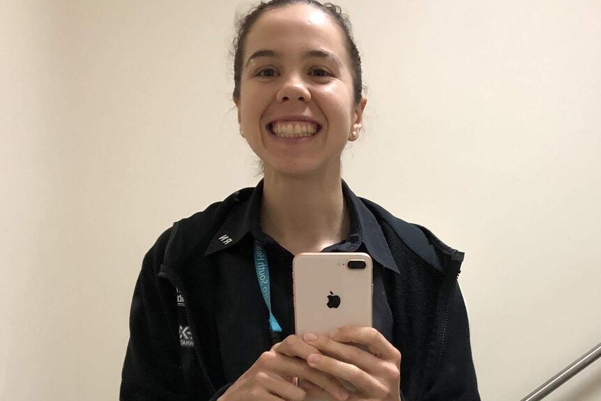 Wearing a nurse's gear and standing in a bathroom, Jess Pratt smiles widely while taking a selfie
