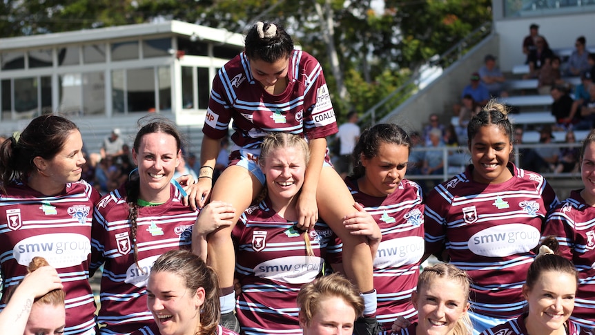A group of women in rugby jerseys celebrate on a football pitch