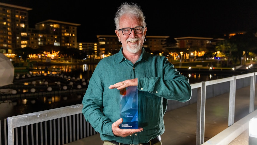 Man with white hair and beard holds blue trophy and faces camera smiling