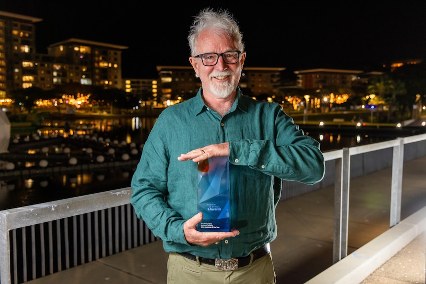 Man with white hair and beard holds blue trophy and faces camera smiling