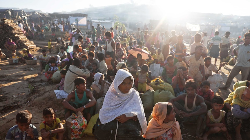 Hundreds of people, some sitting and others standing, under the sun at a camp site.