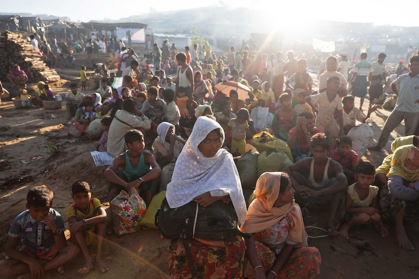 Hundreds of people, some sitting and others standing, under the sun at a camp site.