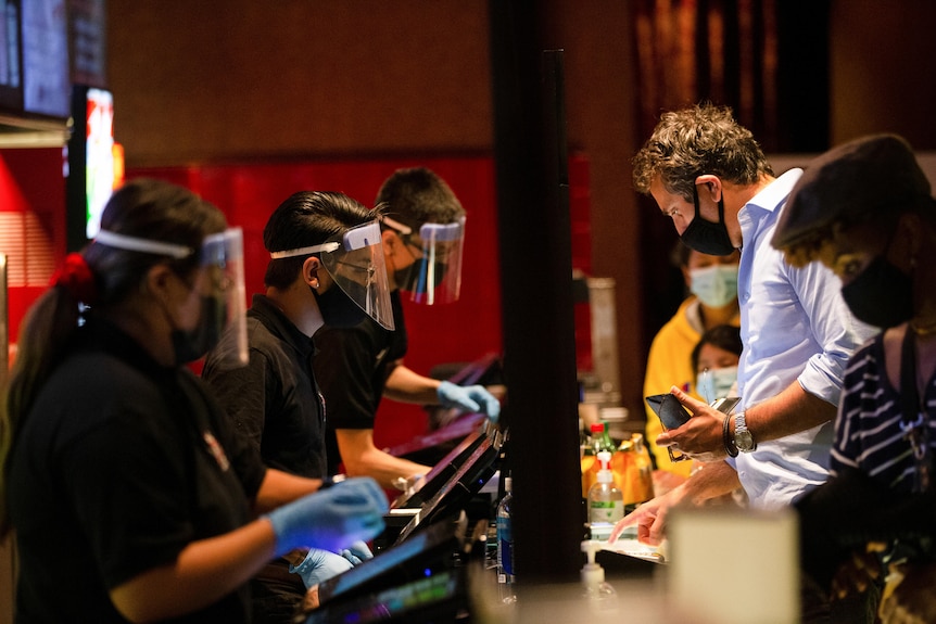 Staff wearing face shield talk to customers behind a stand at a movie theatre.