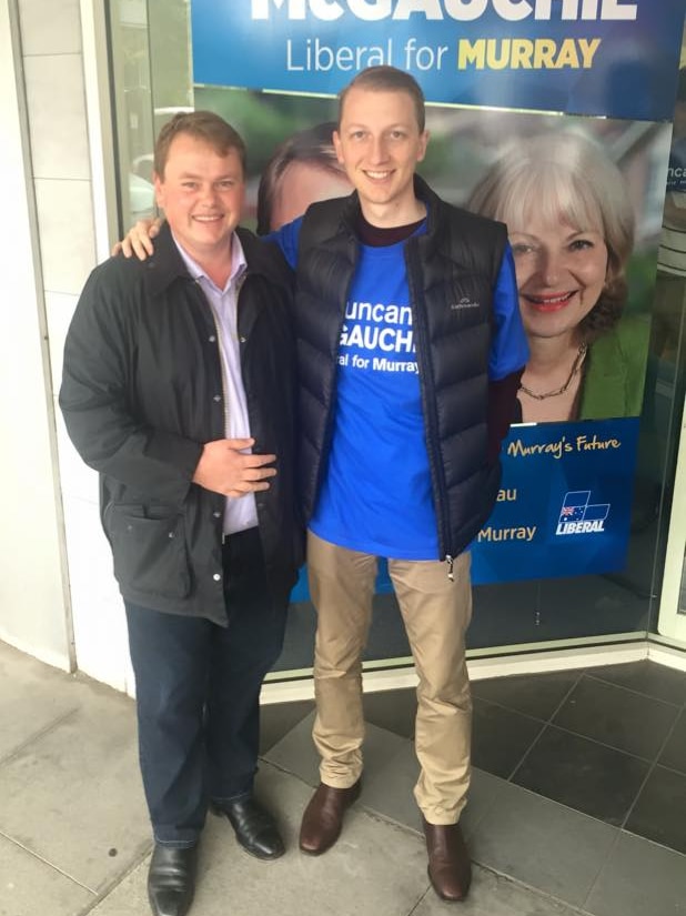 Two men stand in front of a political campaigning sign