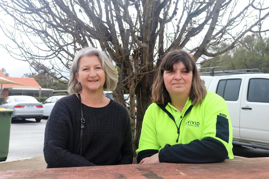 Two women sit at a table facing the camera, smiling. Woman on left is wearing a black shirt, woman on right in fluro work attire