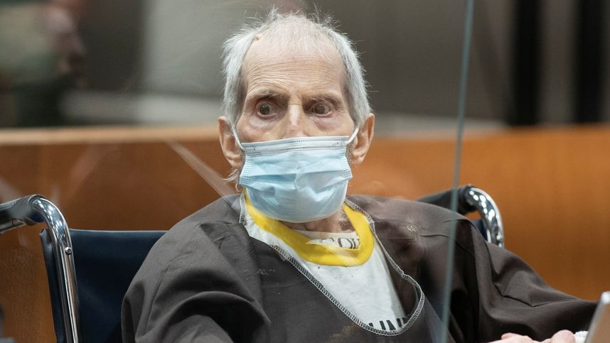 A frail elderly man with grey patchy hair sits in a wheelchair in a court room behind a glass window
