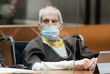 A frail elderly man with grey patchy hair sits in a wheelchair in a court room behind a glass window